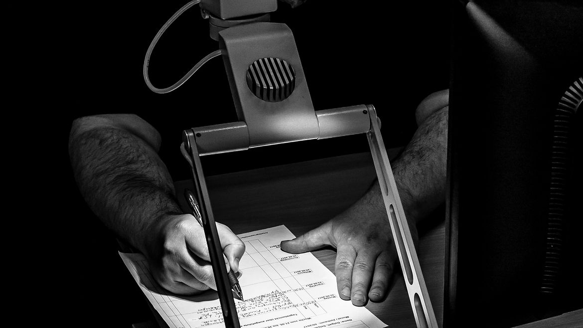 Ein Mann schreibt auf einen Zettel unter einem Lesegerät mit Lampe.