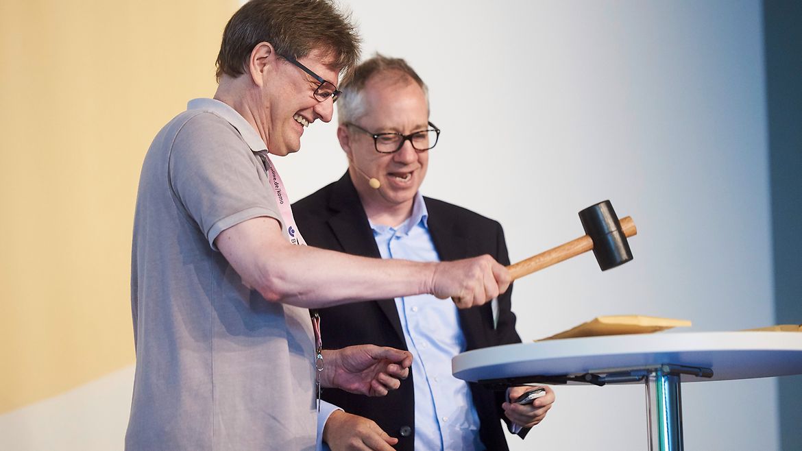 Zwei Männer an einem Stehtisch, auf dem ein Briefumschlag liegt. Einer schlägt mit dem Hammer auf einen Umschlag. Der andere sieht zu. Beide lachen.