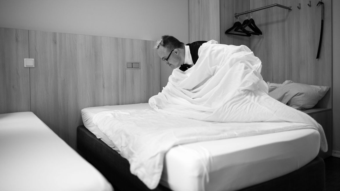 Ein Mann mit Trisomie 21 in einem Hotelzimmer beim Beziehen eines Bettes.