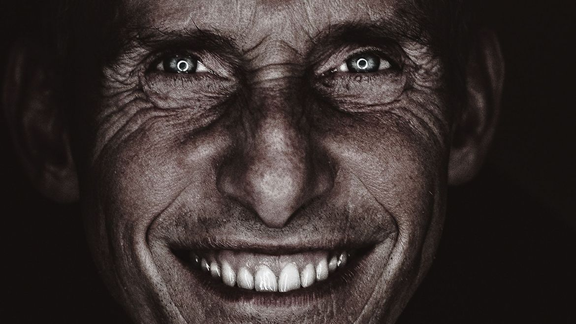 Porträt eines Mannes. Der Hintergrund ist schwarz - das Gesicht wird von vorne beleuchtet. Er lacht in die Kamera und die weißen Zähne leuchten auffällig.