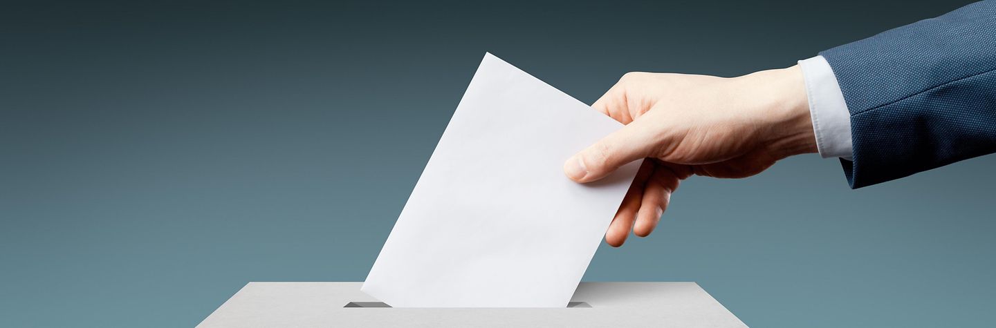 Eine Hand wirft einen Umschlag in den Schlitz einer Wahlurne