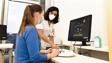 Eine Pflegekraft und eine Patientin in einem Raum mit Computer. Die Patientin hat die Hände auf einer tastaturähnlichen Vorrichtung. Die Pflegekraft gibt Anweisungen