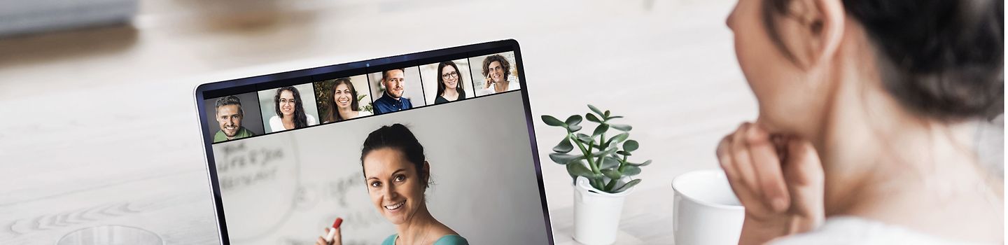 Junge Frau an einem Laptop in einer Videokonferenz - auf dem Bildschirm zeigt eine Frau etwas auf einem Flipchart. Sechs weitere Personen sind am oberen Rand des Bildschirms zu sehen.