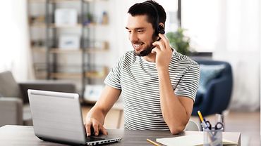 Junger Mann mit Headset sitzt an einem Laptop und spricht in Richtung Bildschirm.