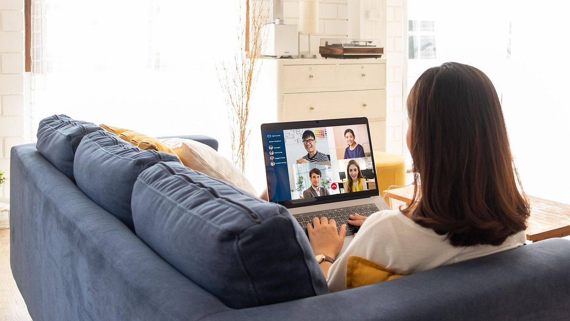 Junge Frau sitzt auf dem Sofa zu Hause. Auf dem Tisch steht ein Notebook - auf dem Bildschirm sind Teilnehmende einer Videokonferenz.