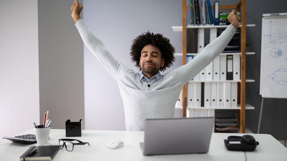 Pausen im Homeoffice Junger Mann arbeitet zu Hause am Computer - er streckt die Arme nach oben.