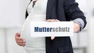 Schwangere Frau in weißer Bluse und dunklem Blazer hält sich mit rechter Hand den Bauch und in der Linken ein Schild mit der Aufschrift Mutterschutz.
