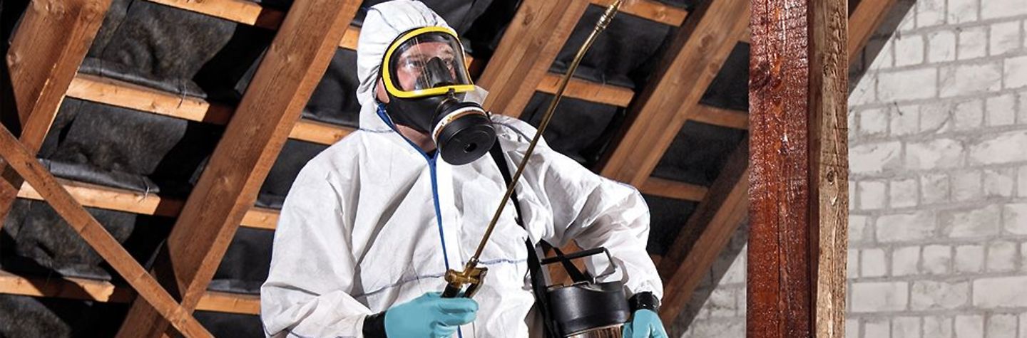 Schädlingsbekämpfer in weißem Schutzanzug mit Gasmaske versprüht ein Mittel auf einem Dachboden