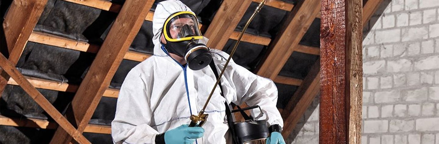 Online-Gefährdungsbeurteilung Schädlingsbekämpfung Schädlingsbekämpfer in weißem Schutzanzug mit Gasmaske versprüht ein Mittel auf einem Dachboden