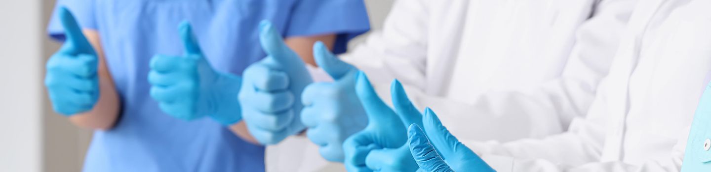 Medizinische Schutzhandschuhe Vier Personen in Kasack bzw. Arztkitteln strecken ihre Hände zusammen nach vorn. Sie tragen blaue medizinische Einmalhandschuhe und heben alle die Daumen.