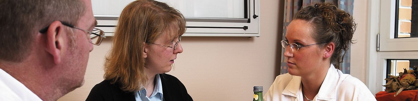 Frau mit dunkler Jacke sowie ein Arzt und eine Ärztin sitzen zum Gespräch an einem Tisch