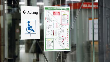 Fluchtpläne Hinweisschild zum Aufzug für Rollstuhlnutzer und Fluchtplan an einer Wand.