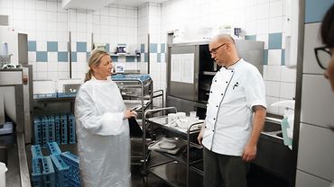 Ruth Giersch spricht mit einem Küchenmitarbeiter neben einem Servierwagen mit Geschirr - sie trägt einen Schutzkittel, er eine Kochjacke.