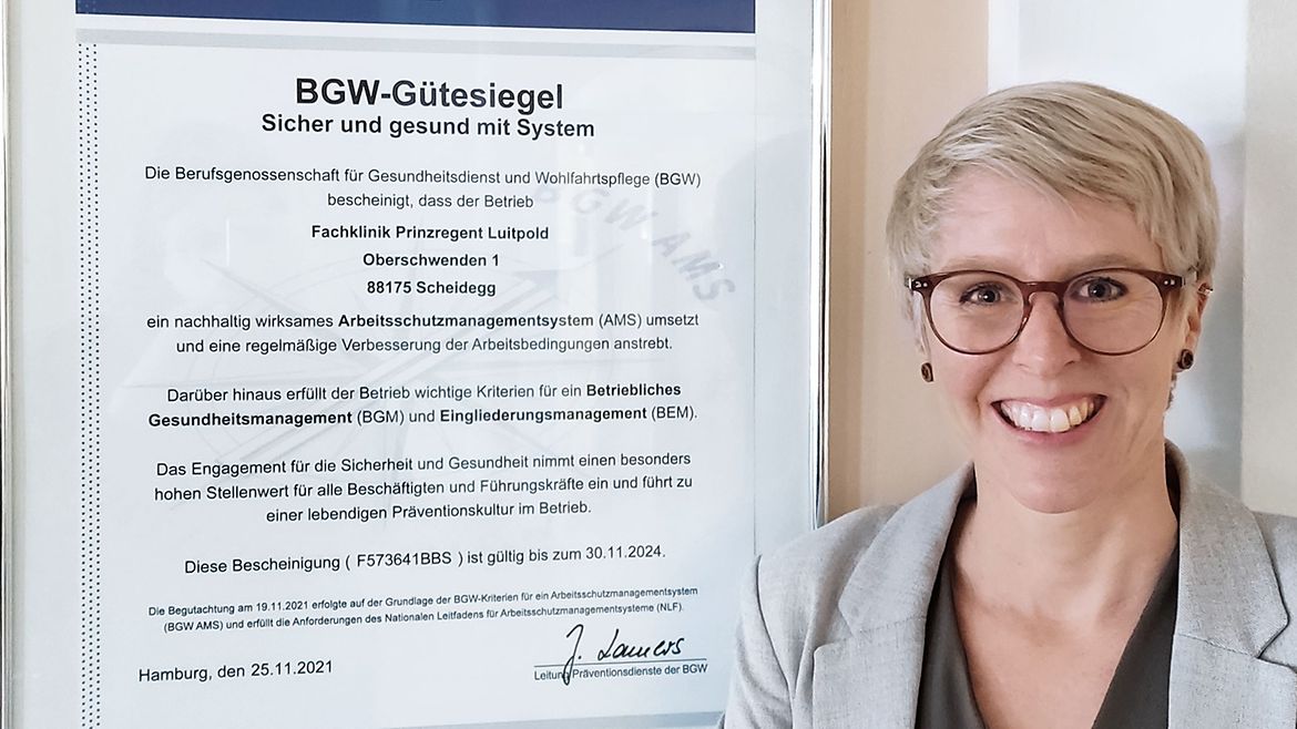Arbeitsschutzmanagement Julia Schäfer lacht in die Kamera. Hinter ihr hängt eine Urkunde des BGW-Gütesiegels an der Wand.