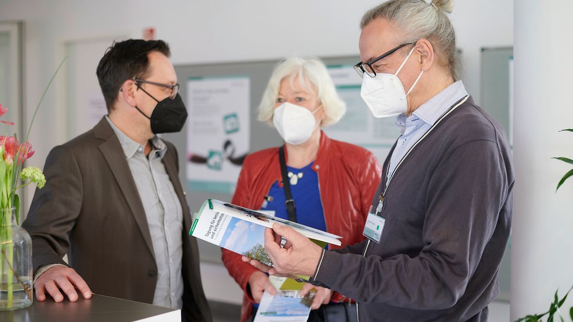 Drei Personen stehen am Anmelde-Counter - zwei blättern in einer Broschüre.