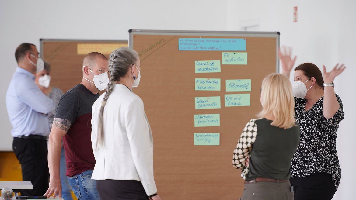 Fünf Teilnehmende stehen an einer Pinwand in einem Workshop. Alle tragen einen Mund-Nasen-Schutz.