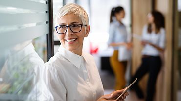 Eine Unternehmerin lehnt lächelnd mit einem iPad in der Hand an einer Glaswand. Im Hintergrund unterhalten sich 2 Beschäftigte.