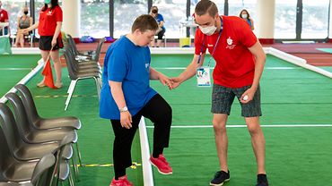 BGW-Volunteer im roten Trikotshirt hilft einer Boccia-Spielerin im blauen Shirt auf die Spielfläche