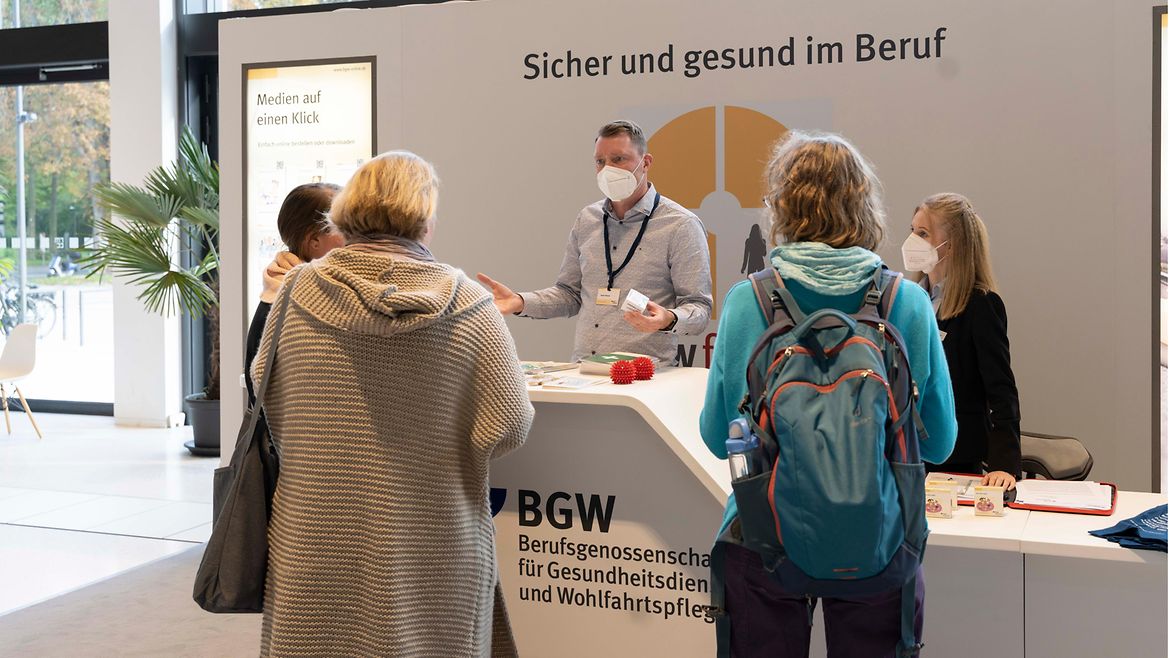 Mehrere Personen stehen am einen Messestand, ein Mann mit Maske hinter dem Tresen spricht mit ihnen, im Hintergrund steht an der Rückwand der Text "Sicher und gesund im Beruf"
