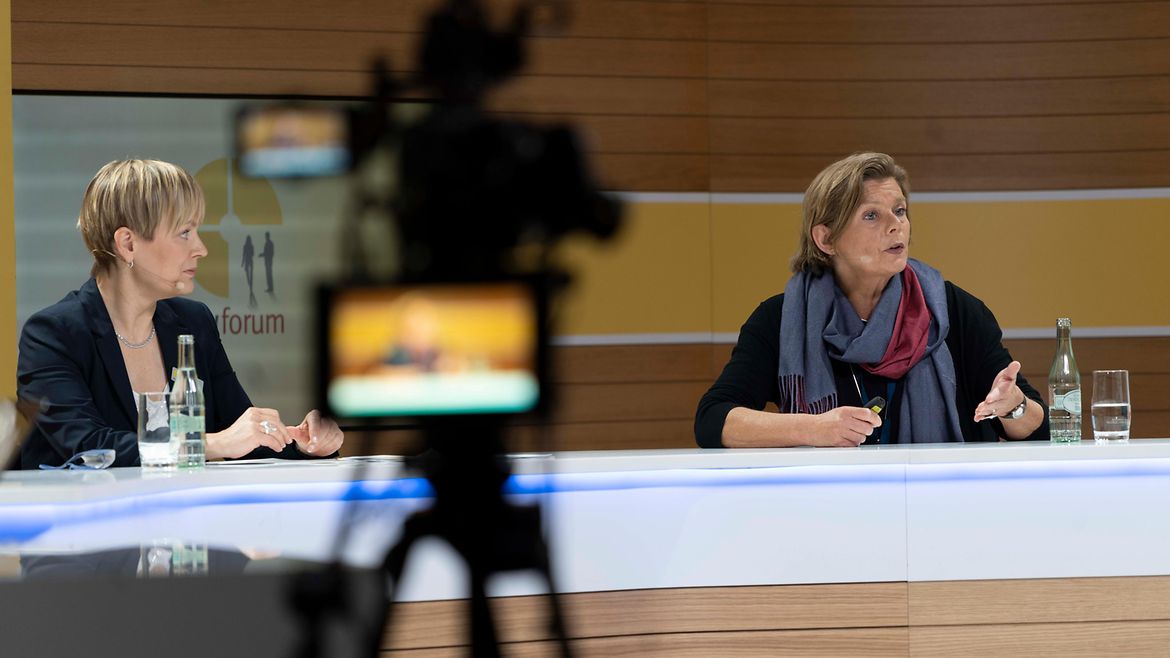 Zwei Frauen sitzen an einem Tisch wie in einem Fernsehstudio, im Bildvordergrund eine verschwommene Kamera, durch die sie gefilmt werden