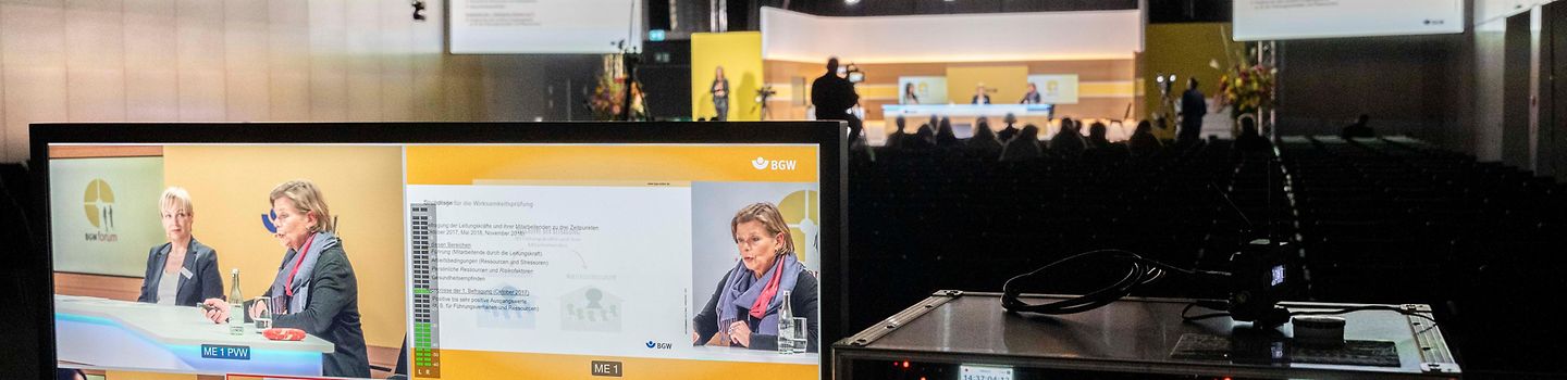 BGW forum 2022 - Technik Mischpult im Bildvordergrund mit vielen verschiedenen Kameransichten, im Bildhintergrund gut ausgeleuchtetes Podium mit einer Diskussionsrunde und Kameramann