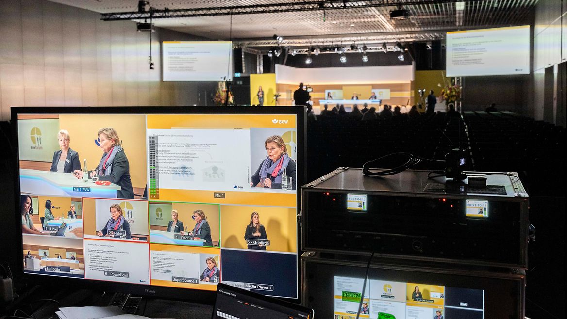 Mischpult im Bildvordergrund mit vielen verschiedenen Kameransichten, im Bildhintergrund gut ausgeleuchtetes Podium mit einer Diskussionsrunde und Kameramann