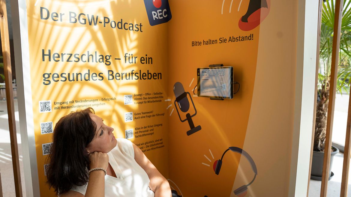 Eine Frau sitzt vor einer Wand mit dem Text "Der BGW-Podcast" und "Herzschlag - für ein gesundes Berufsleben".
