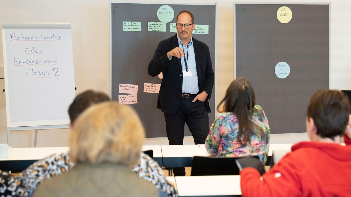 Ein Mann steht in einem Seminarraum vor Teilnehmenden. Hinter ihm befinden sich zwei Pinwände mit Karten, neben ihm steht ein Flipchart mit der Aufschrift: "Beherrschendes oder beherrschtes Chaos?"