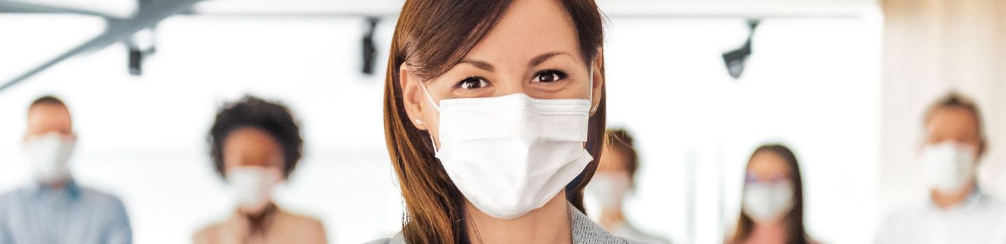 Gemeinsam zum Erfolg Eine Frau schaut direkt in die Kamera. Hinter ihr stehen weitere Menschen, die ebenfalls nach vorn schauen. Alle tragen Masken.