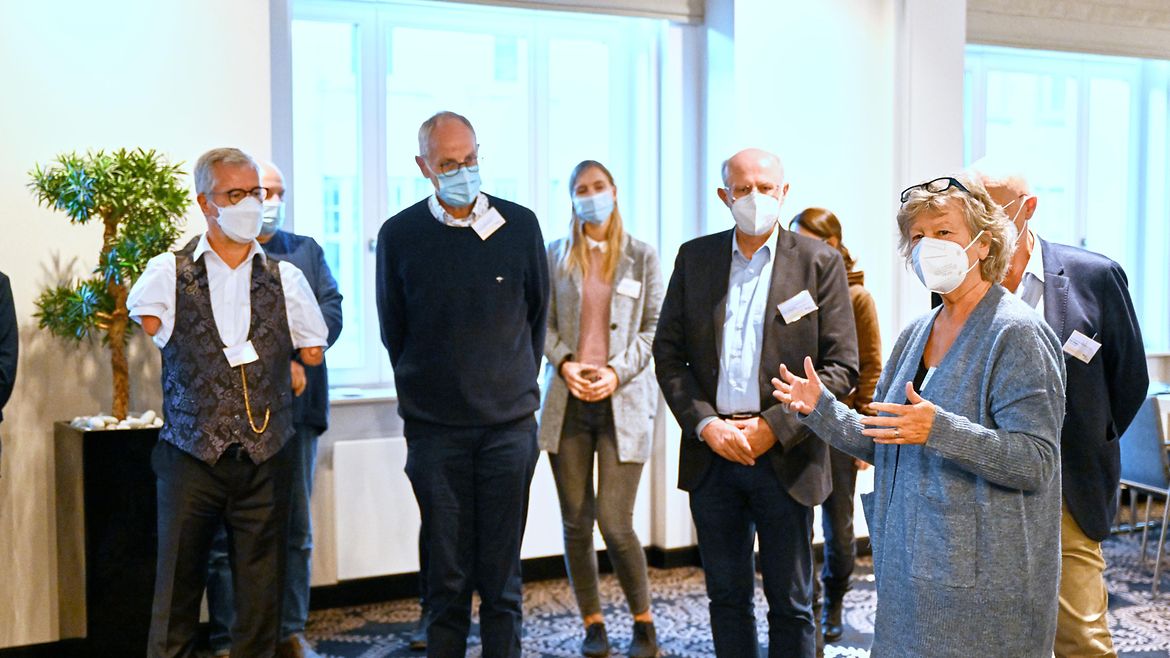 Eine Gruppe Personen steht in einem Raum und spricht miteiander - ganz links: Moderatur Rainer Schmidt. Alle tragen FFP-2-Masken.