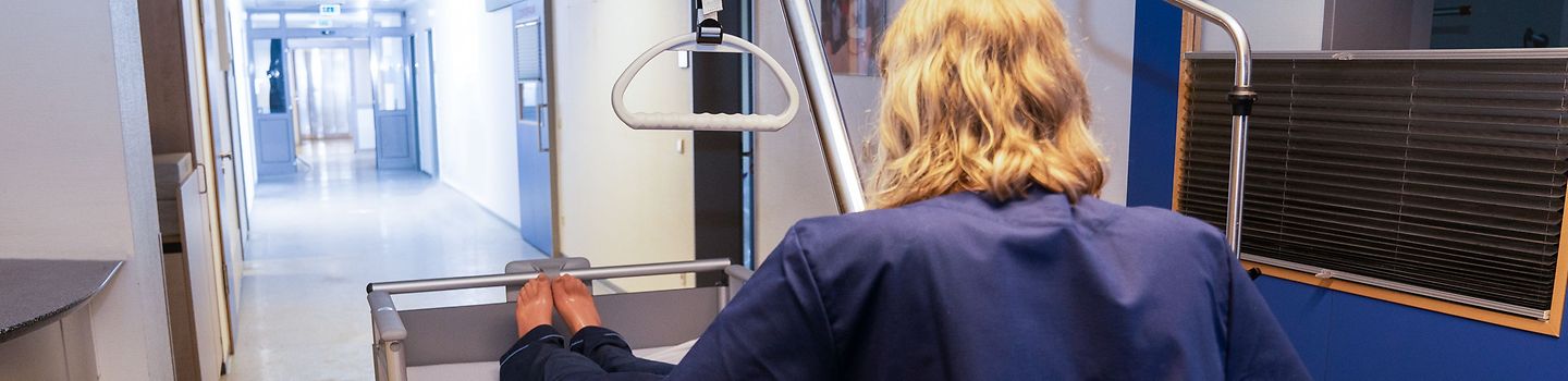 BGW test 2022 - Krankenhausbetten Eine Pflegeperson schiebt ein Pflegebett mit Patienten-Dummy über einen Flur.
