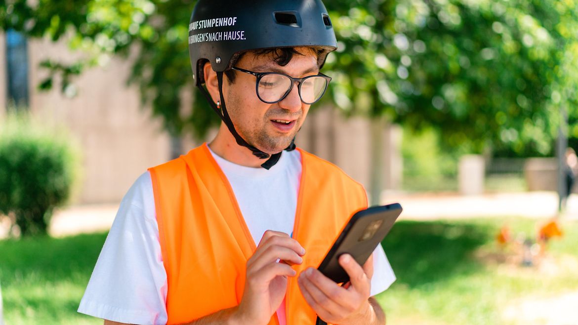 Junger Mann mit Warnweste und Helm schaut auf ein Smartphone und tippt etwas ein.