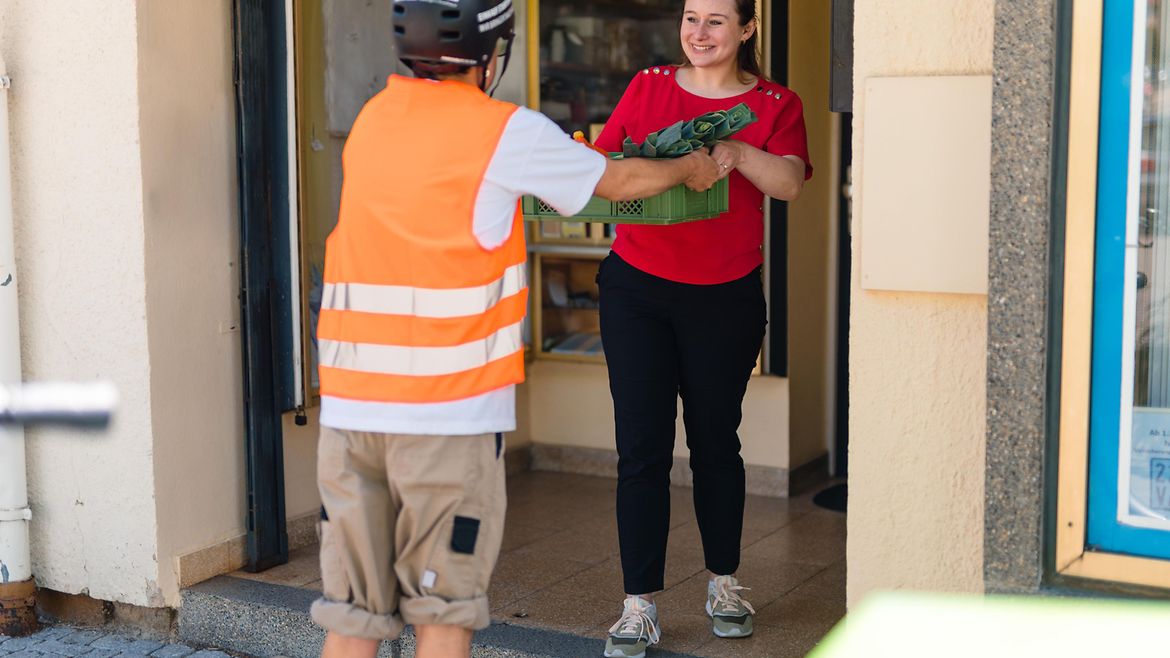 Junger Mann mit Warnweste, Helm und kurzen Hosen liefert eine Kiste mit Gemüse an eine Haustür. Eine junge Frau nimmt die Kiste entgegen.