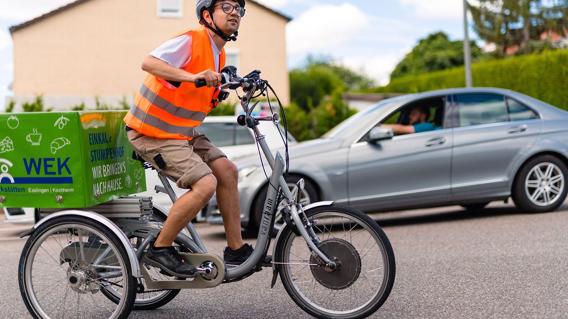 Junger Mann mit Warnweste, Helm und kurzen Hosen fährt auf einem Lastenrad die Straße entlang. Ein Auto fährt in die entgegengesetzte Richtung