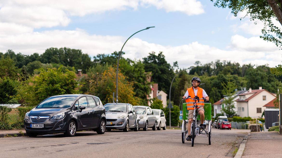 Junger Mann mit Warnweste, Helm und kurzen Hosen fährt auf einem Lastenrad eine Straße entlang - hinten auf dem Rad befindet sich ein großer Behälter zum Transport von Waren. Auf der rechten Seite der Straße parken Autos.