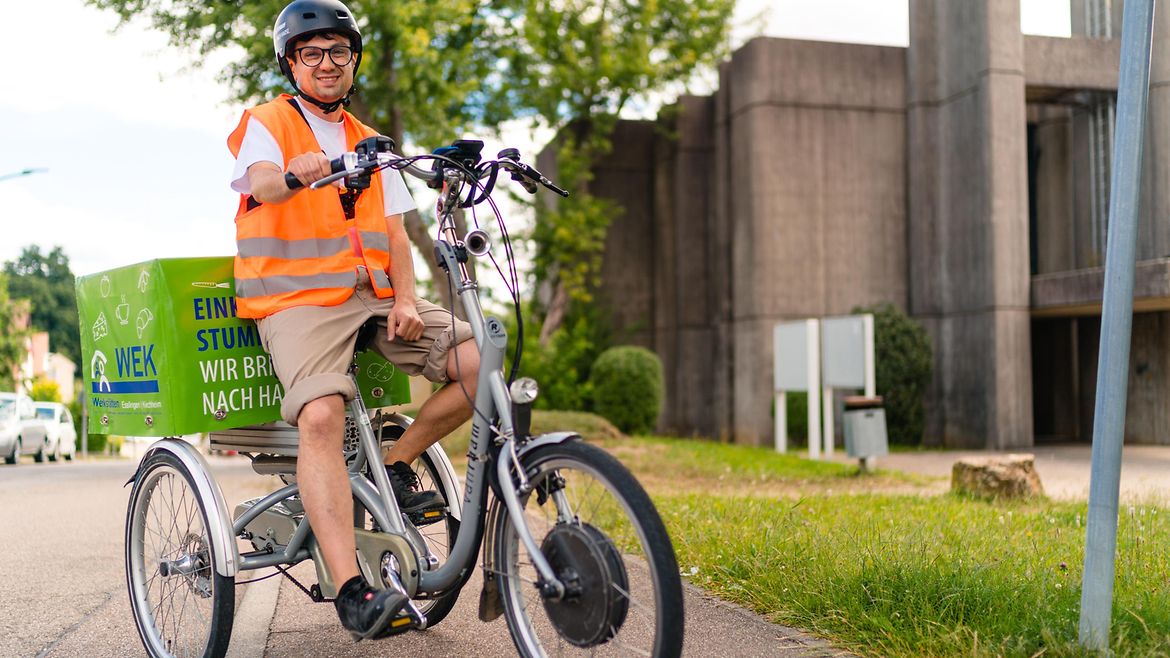 Junger Mann mit Warnweste, Helm und kurzen Hosen sitzt auf einem Lastenrad - hinten auf dem Rad befindet sich ein großer Behälter zum Transport von Waren.