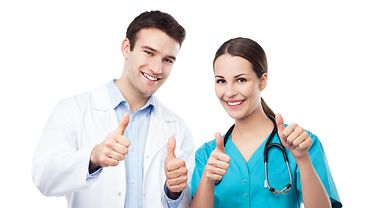 Zwei Personen lachen in die Kamera und zeigen mit den Daumen nach oben - beide tragen Pflege- oder Arzt-Berufskleidung. Eine Person trägt ein Stethoskop um den Hals.