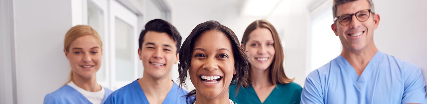 Gruppe von 5 Personen in einem Krankenhausflur. Alle tragen Krankenhaus-Berufskleidung und lachen in die Kamera.