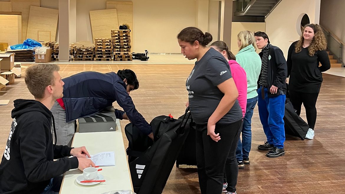 Fünf Frauen stehen in einer Schlange vor einem Tisch, hinter dem zwei Volunteers Sport-Equipement austeilen.