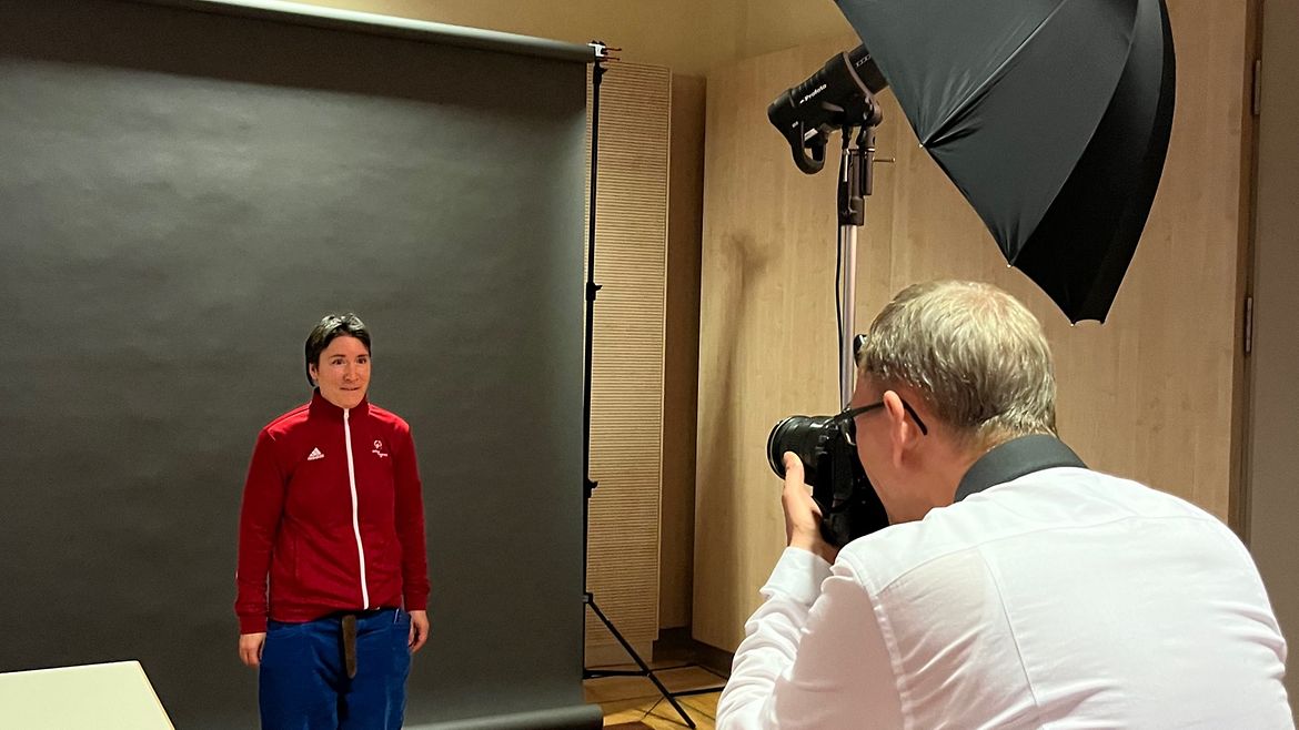 Eine Frau steht in roter Trainingsjacke vor einem schwarzen Hintergrund. Im Vordergrund der Rücken des Fotografen mit Kamera.