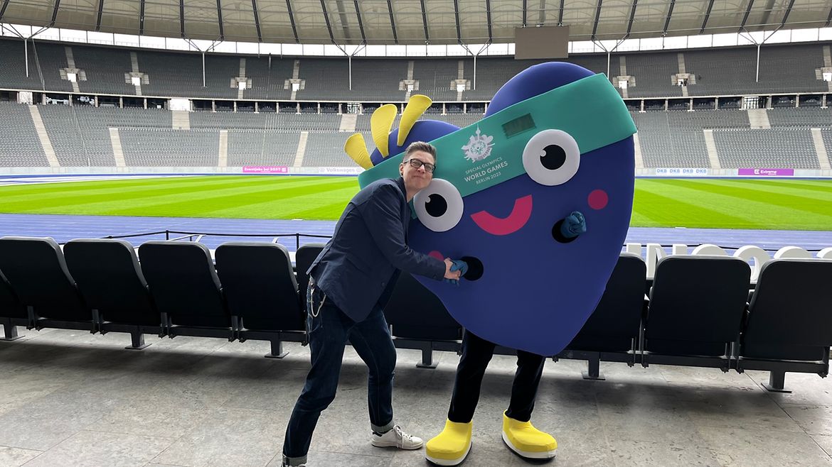 Eine Frau steht im Olympiastadion Berlin neben dem Maskottchen Unity - ein verkleideter Mensch -und gibt ihm die Hand.