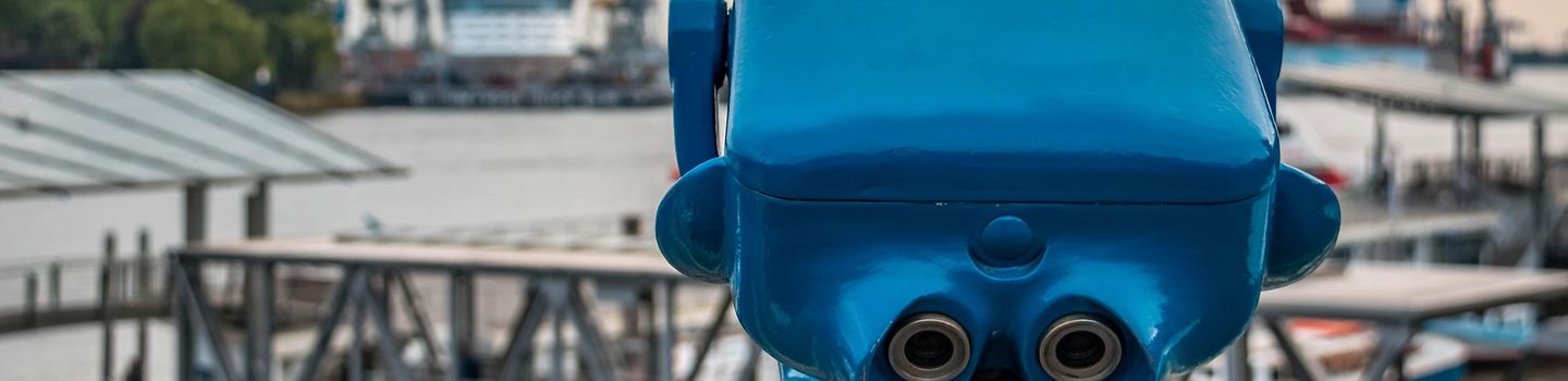 Hamburger Gesundheitspreis Hamburg Landungsbrücken: Vorn ein großes blaues Fernglas, Blick auf den Hafen mit Kränen und Pontonbrücken. Der Himmel ist bewölkt.