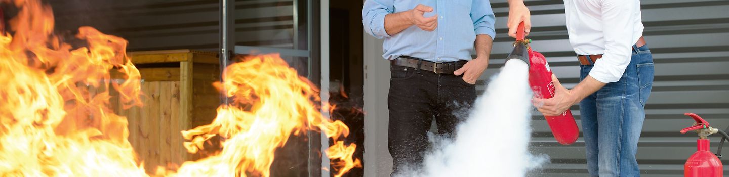 Zwei Männer im Freien vor einem Gebäude. Einer löscht einen Brand mit einem Feuerlöscher, der andere steht neben ihm und erklärt etwas. Neben ihnen stehen weitere Feuerlöscher.