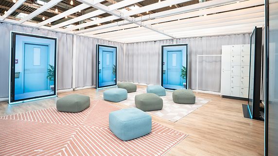 Blick in die Ausstellung "In Würde Abschied nehmen": Grüne und blaue Sitzhocker sind im Raum verstreut, an den Wänden vier menschenhohe Bildschirme, die eine Tür zeigen.
