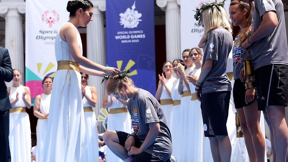 Die Hohepriesterin im weißen, antiken Gewand legt die Hände auf den Kopf der Special Olympics Athletin Natascha Wermelskirchen, die vor ihr kniet. Daneben weitere Teilnehmende, im Hintergrund Frauen in weißen Gewändern.