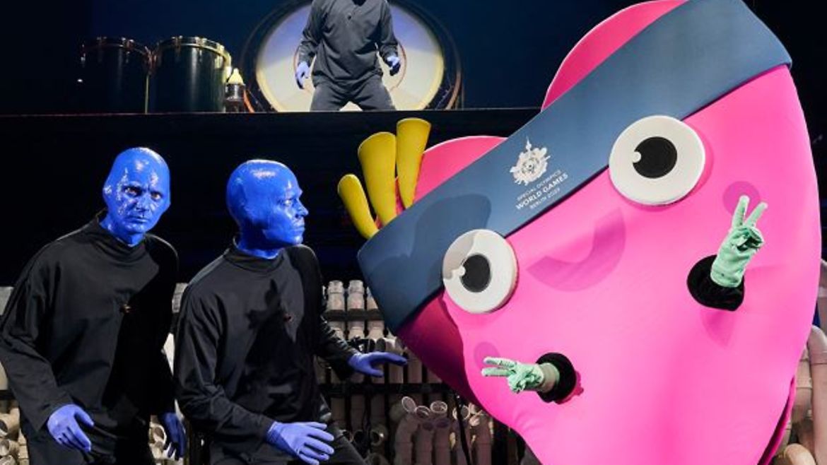 Das Maskottchen Unity - ein Plüschherz, in dem ein Mensch steckt - mit zwei blauen angemalten Menschen auf einer Bühne