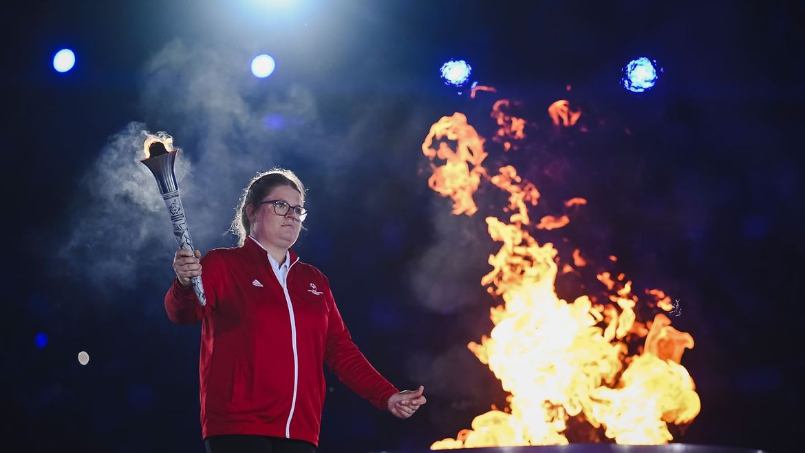 Eine Athletin in roter Trainingsjacke mit einer brennenden Fackel in der Hand steht vor dem entzündeten Feuer.