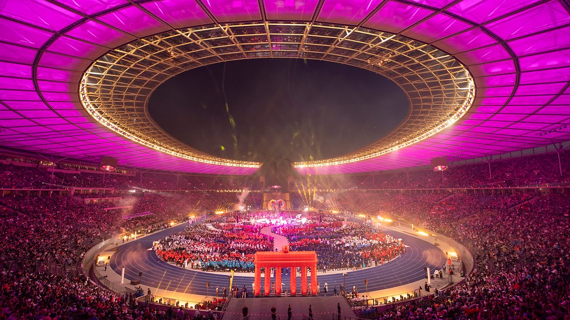 Das Olympiastadion in violettes Licht getaucht. In der Mitte stehen alle Delegationen zusammen. Davor ist ein aufblasbares Brandenburger Tor zu sehen.