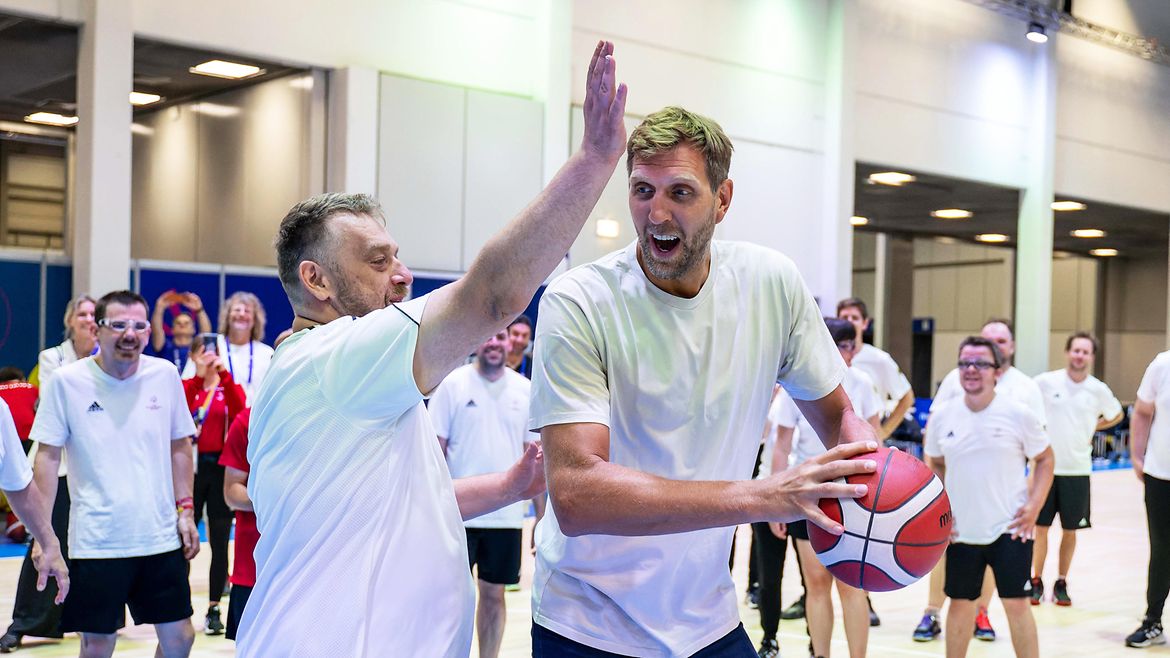 Dirk Nowitzki neben Marco Klein in einer Halle. Nowitzki hält einen Basketball, Klein hebt den Arm zur Abwehr. Im Hintergrund weitere Sportler.