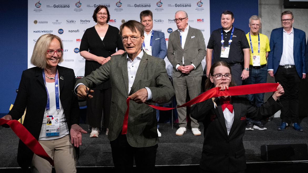 Christiane Krajewski neben Karl Lauterbach und Hannah Atkinso Joy: Lauterbach hält ein Schere, mit der er das rote Band daurchschnitten hat, das alle drei halten. Im Hintergrund 6 Menschen